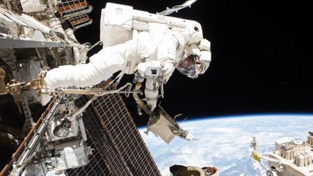 An astronaut in a white space suit faces toward the bottom of the image amidst a series of space equipment floating above Earth. 
