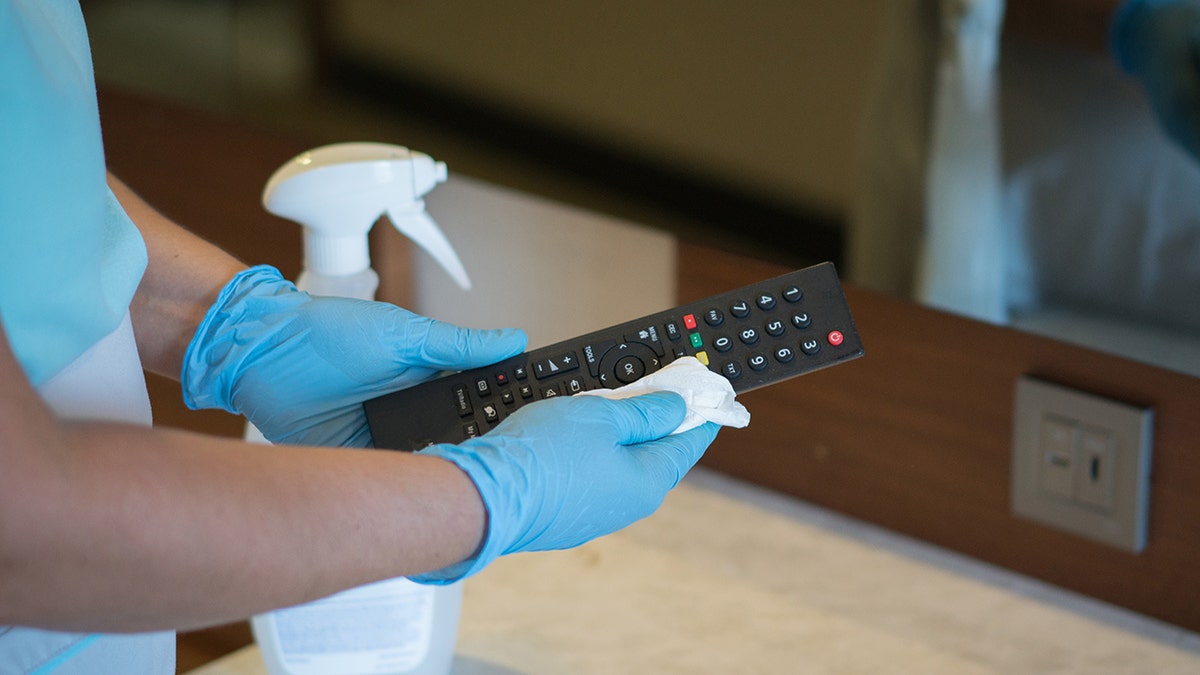 A housekeeping staffer wiping down a remove in a hotel room, seen wearing blue gloves with spray bottle behind her
